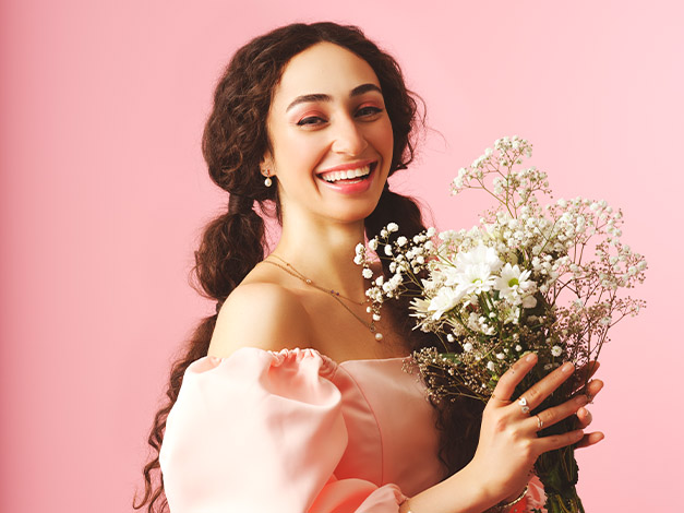 Frau mit langem lockigem Haar in rosa Kleid lächelt und hält einen Strauß weißer Blumen vor rosa Hintergrund.
