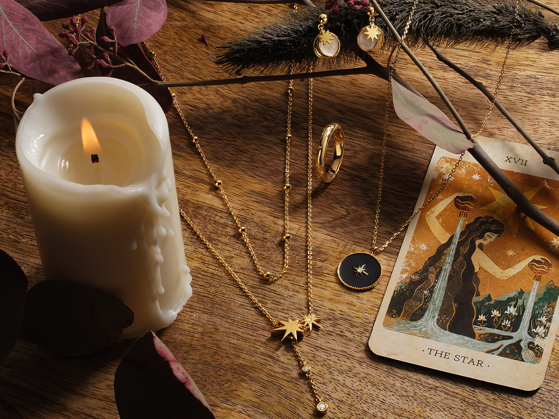 Goldschmuck mit Sternmotiven auf Holztisch, daneben brennende Kerze und Tarotkarte ‚The Star‘.
