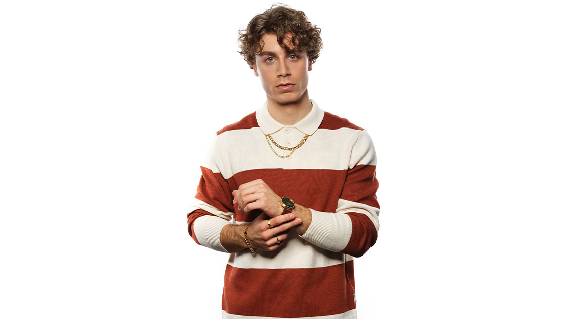 Junger Mann mit lockigem Haar trägt gestreiften Pullover in Weiß und Rostrot sowie goldene Ketten und Armbanduhr.