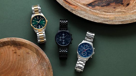 Drei FAVS Herrenarmbanduhren auf dunkelgrünem Grund mit zwei Holztellern als Deko-Elementen.